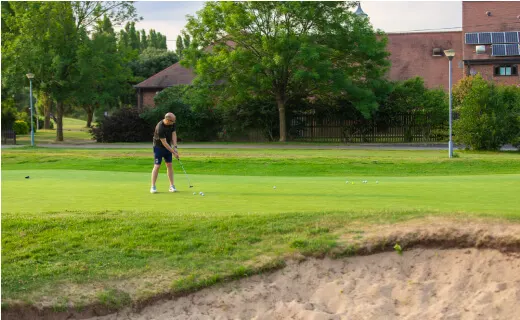 Golfer playing a shot on a short par-3 hole with a sand bunker in the foreground.