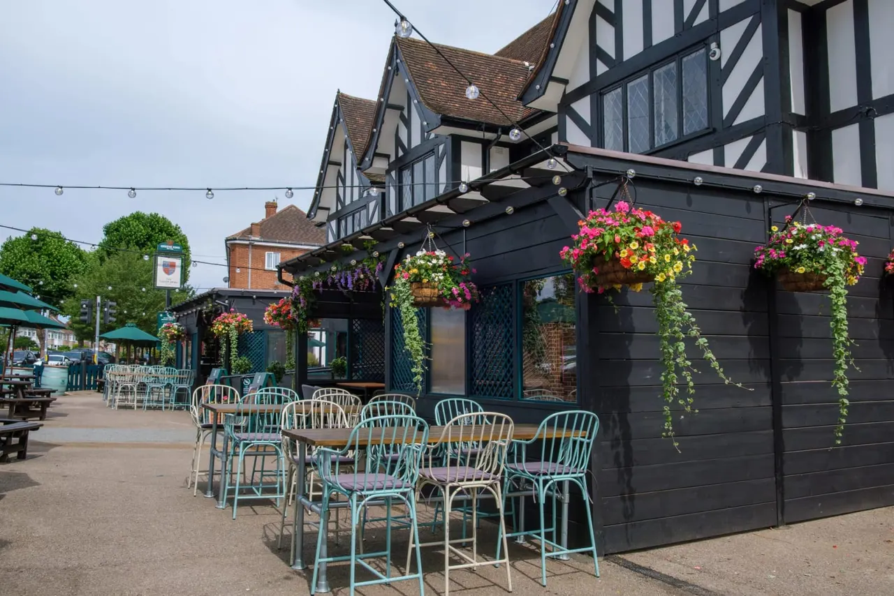 Outdoor seating area with turquoise bar stools and chairs beneath a black pergola decorated with hanging baskets of pink, yellow and purple flowers.