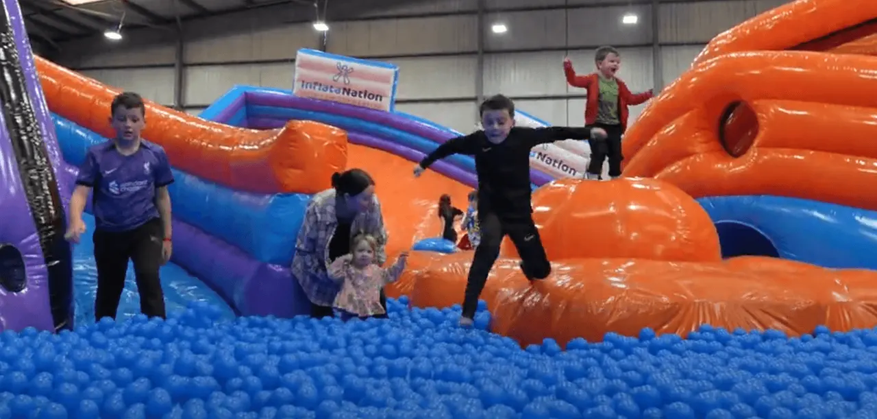 Children and adults playing in a large indoor inflatable park with orange and blue bouncy structures and a ball pit.