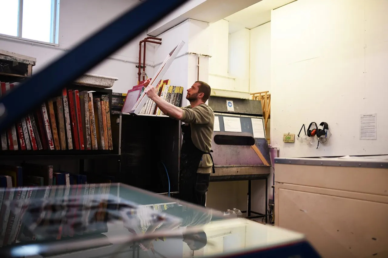 A man reaches up to place a book on a shelf in a workshop room lined with shelves of books and equipment.