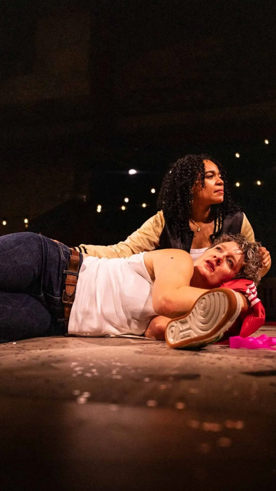 Two actors perform an intimate scene on stage, one lying down whilst the other leans over them.