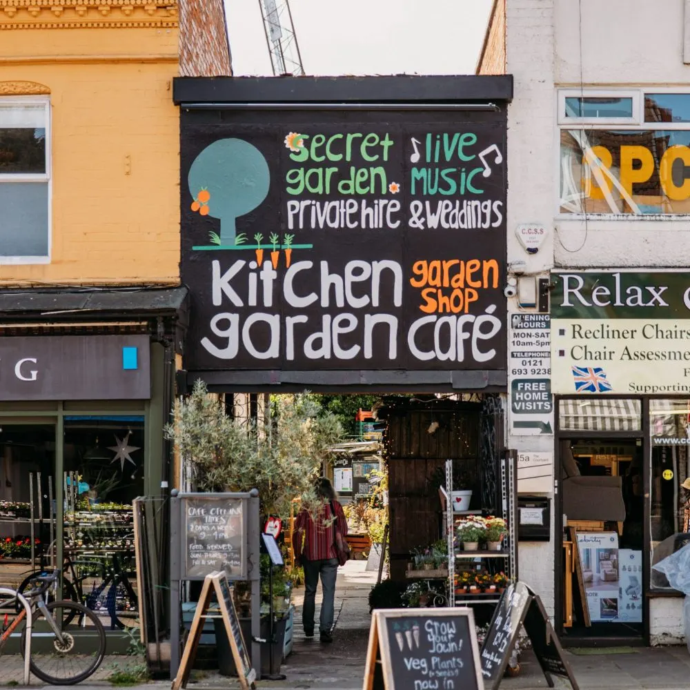 Street-level view of a café storefront with hand-painted signage advertising a secret garden, live music, kitchen garden, and café facilities.