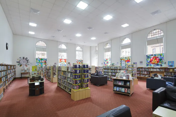 Bright, spacious library interior with bookshelves, colourful artwork on tall windows, red carpeting, and seating areas beneath arched windows.