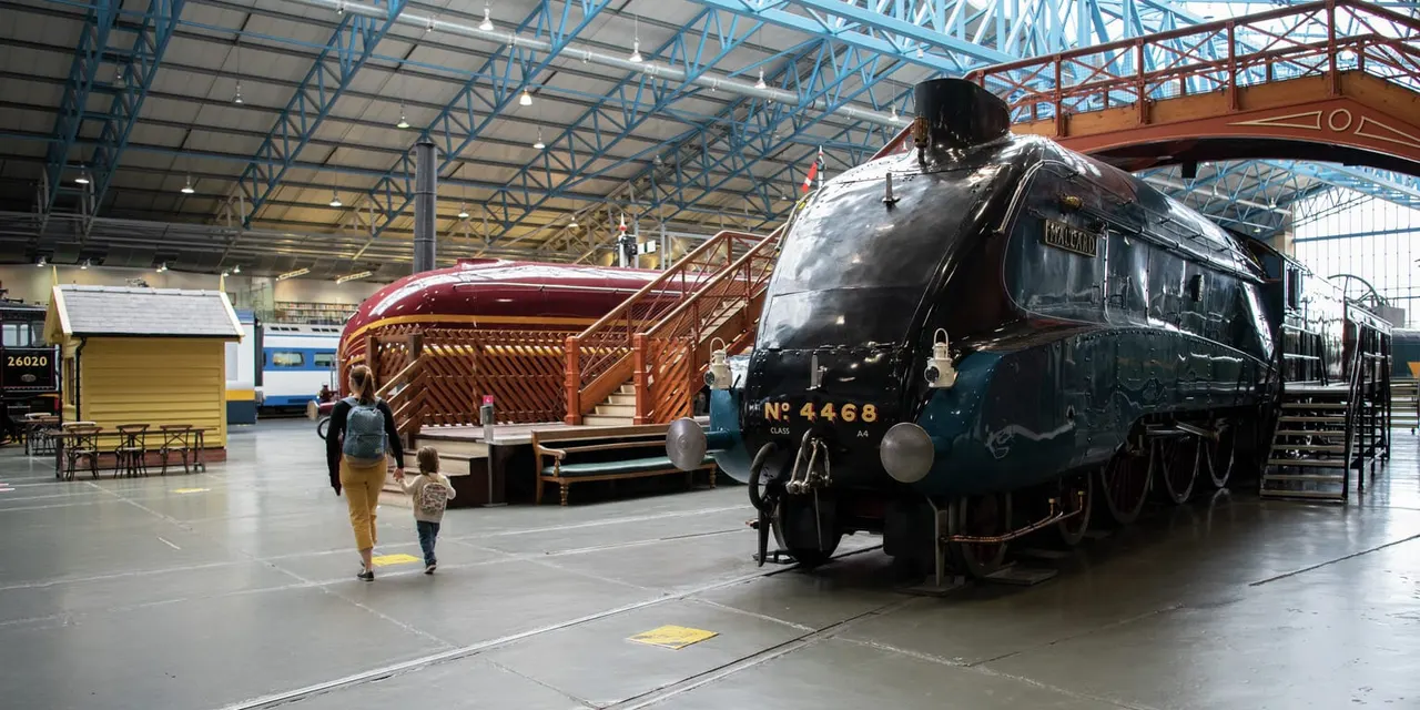 Adult and child walking past a vintage steam locomotive and other heritage trains in a large railway depot hall.