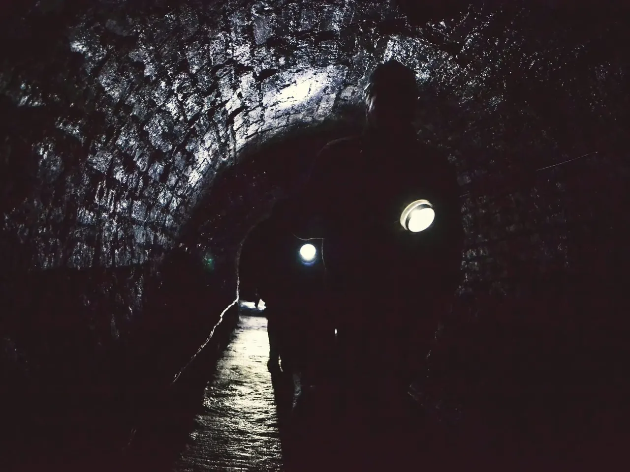 A person walks through a dark stone tunnel lit by electric lamps, with water on the floor and openings in the ceiling showing light above.