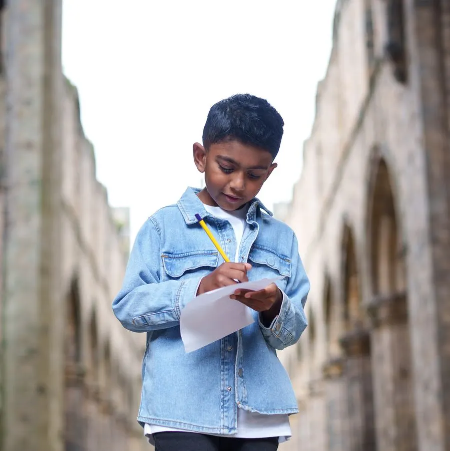 Young child in denim shirt writing on clipboard between stone walls of historic abbey ruins.