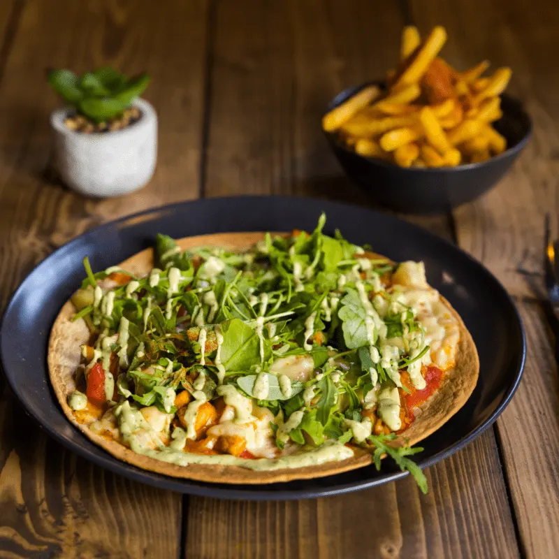Pizza topped with cheese, vegetables and fresh salad leaves, served with chips and a succulent plant on wooden table.