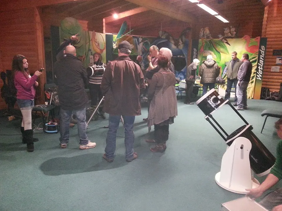 A group of visitors gathered indoors at an astronomy venue, with telescopes and educational displays visible around the room.