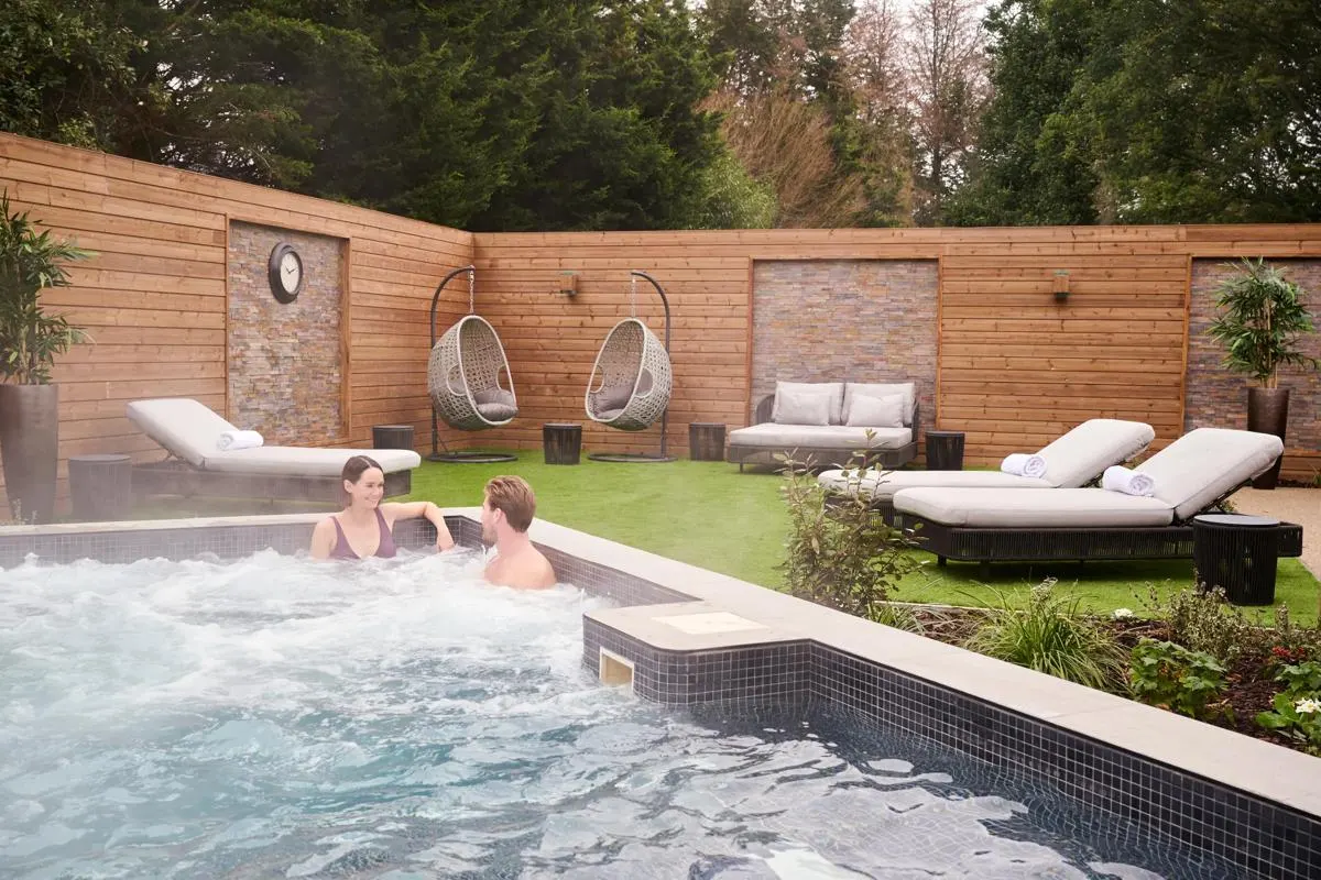 Two people enjoying a hot tub in a spa garden with loungers, hanging chairs and wooden fencing.