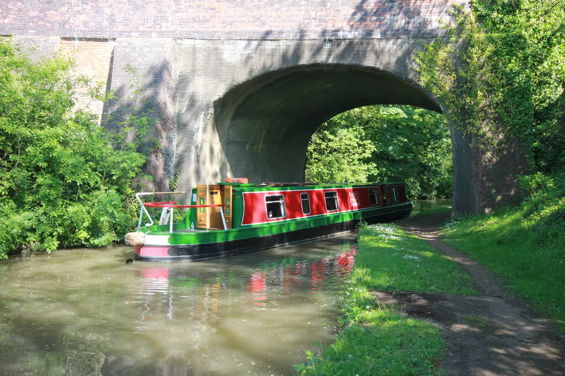 Colourful narrowboat moored on a canal approaching a brick railway arch bridge, surrounded by green vegetation.