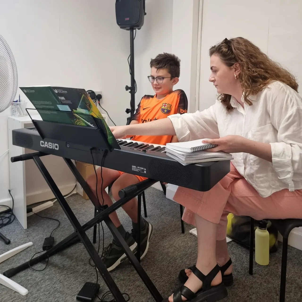 An instructor teaches a young boy piano during a lesson in a music studio with a speaker and microphone.