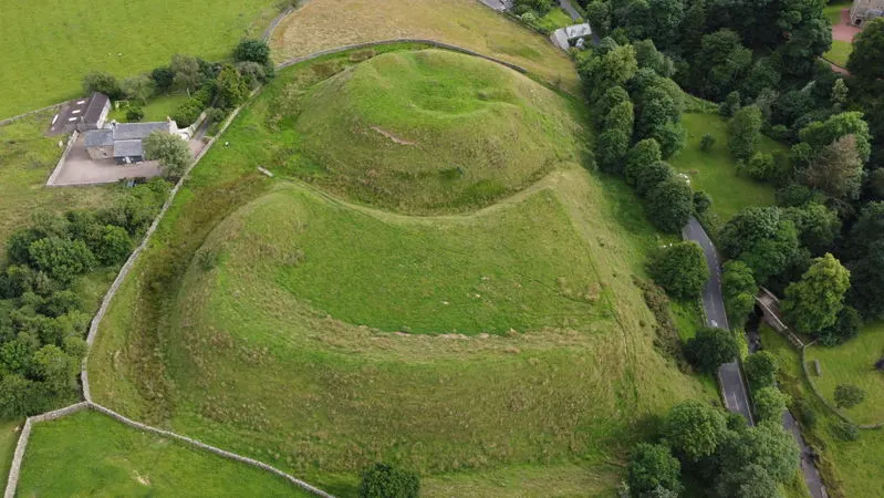 Aerial view of ancient earthwork mounds surrounded by green fields and woodland pathways.