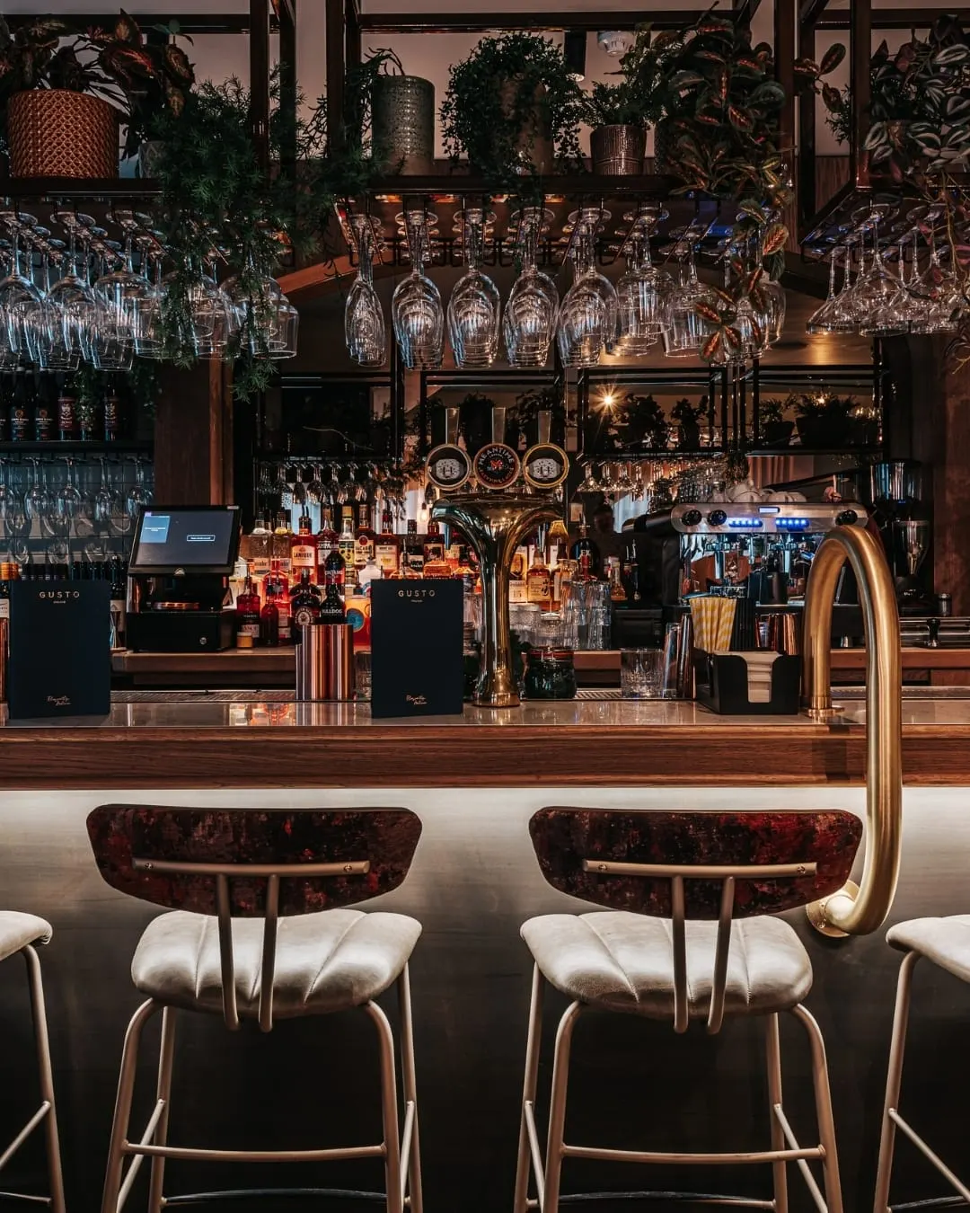 Bar counter with upholstered stools, shelves of spirits and hanging glassware, potted plants and warm lighting overhead.