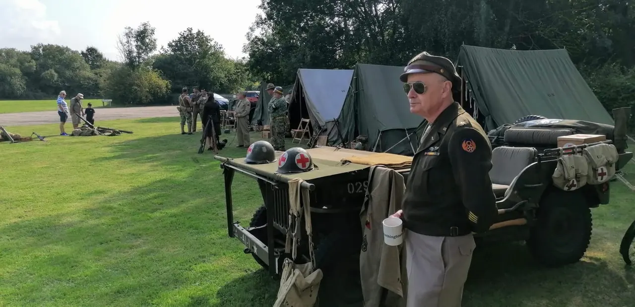 WWII military re-enactor in uniform standing beside vintage military vehicle and equipment on grass field with canvas tents and other participants in background.