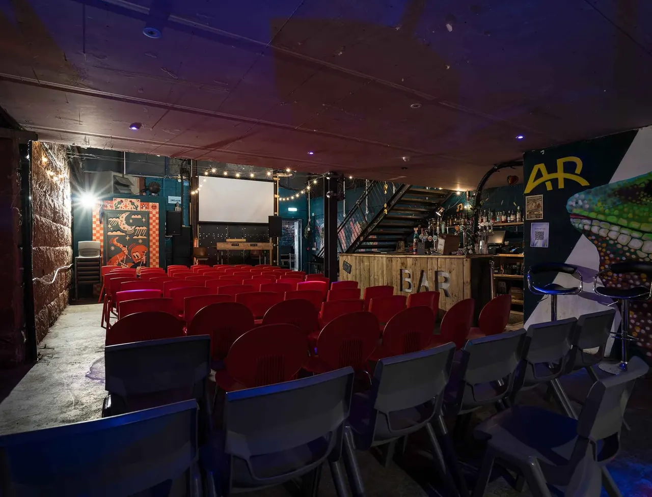 Cinema screen room with rows of red seats facing a blank projection screen, bar counter to the right, and atmospheric lighting throughout the indoor venue.
