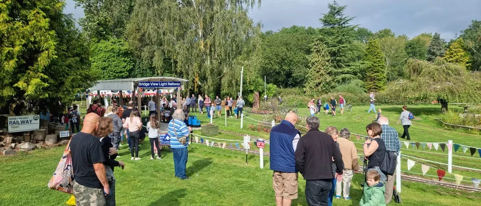Outdoor garden party or community event with visitors strolling on green lawns between white-fenced enclosures, with trees and a railway building in background.