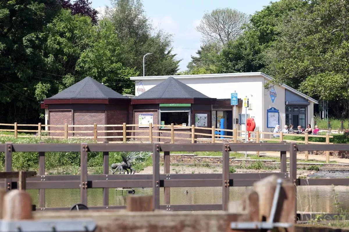 A waterside building with brick and white sections surrounded by trees, viewed across a wooden railing beside a canal.