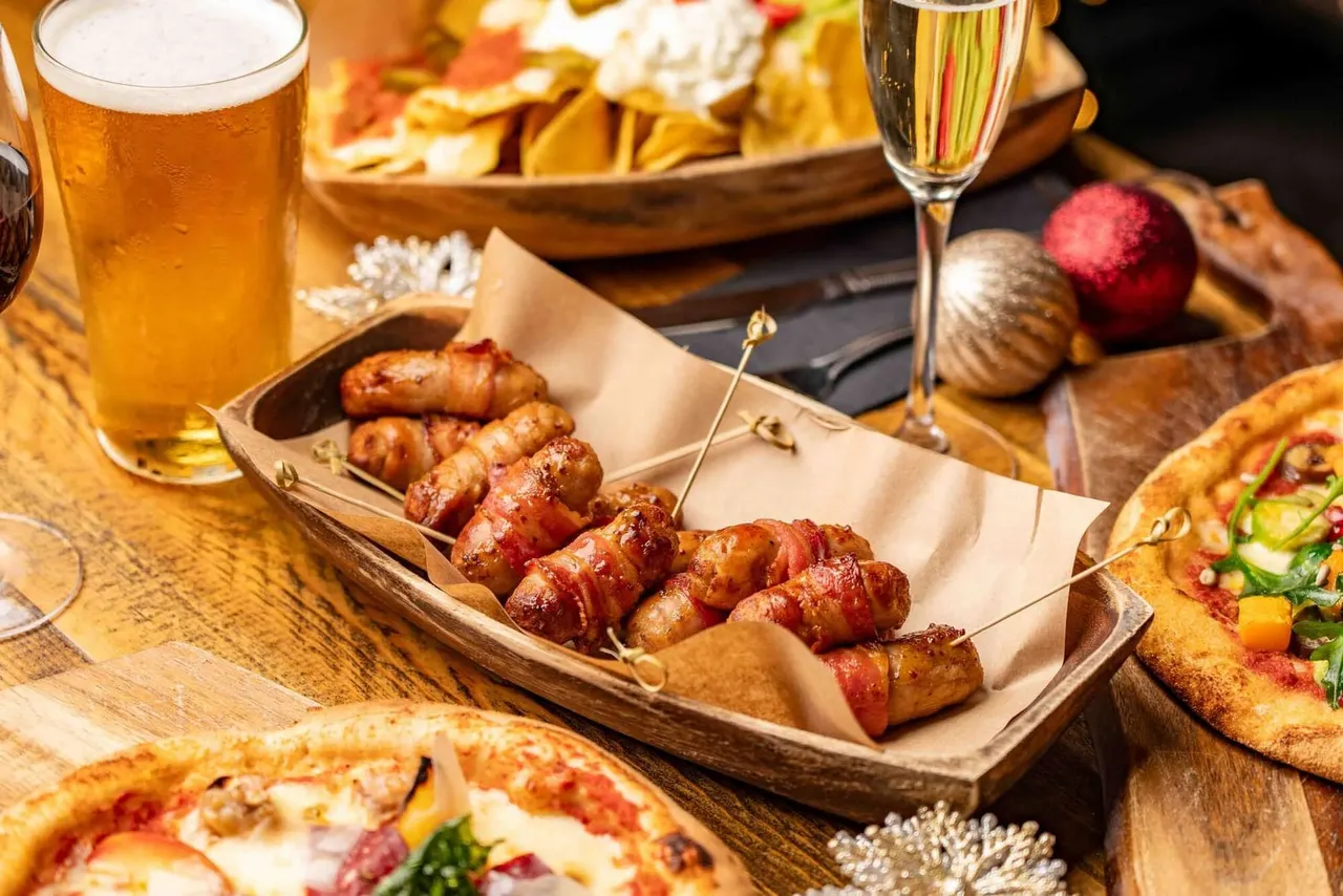 Festive table setting with bacon-wrapped sausages, pizzas, chips, beer, champagne and Christmas decorations.