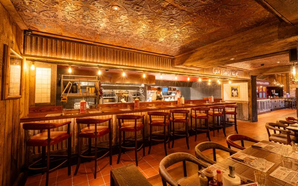 Interior of a restaurant with wooden bar seating, open kitchen visible behind counter, and dining tables in foreground.