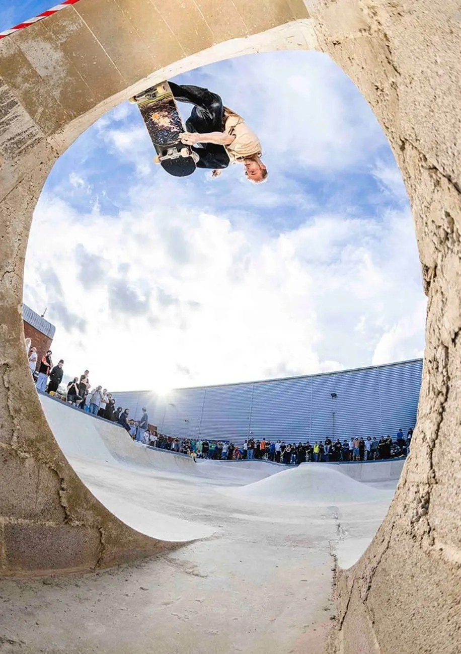 Skateboarder performing an inverted aerial trick above a concrete bowl while spectators watch from the park's edge.