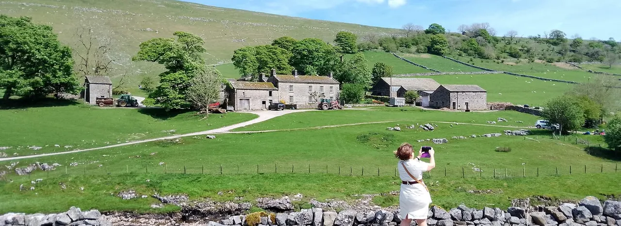 Person in white dress photographing a rural stone farmstead nestled in green rolling hills with dry stone walls and scattered trees.