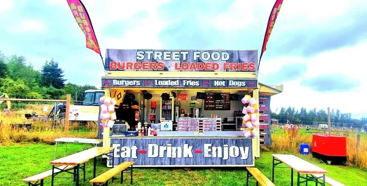 A colourful street food van with picnic benches in a rural outdoor setting with green fields and trees behind.