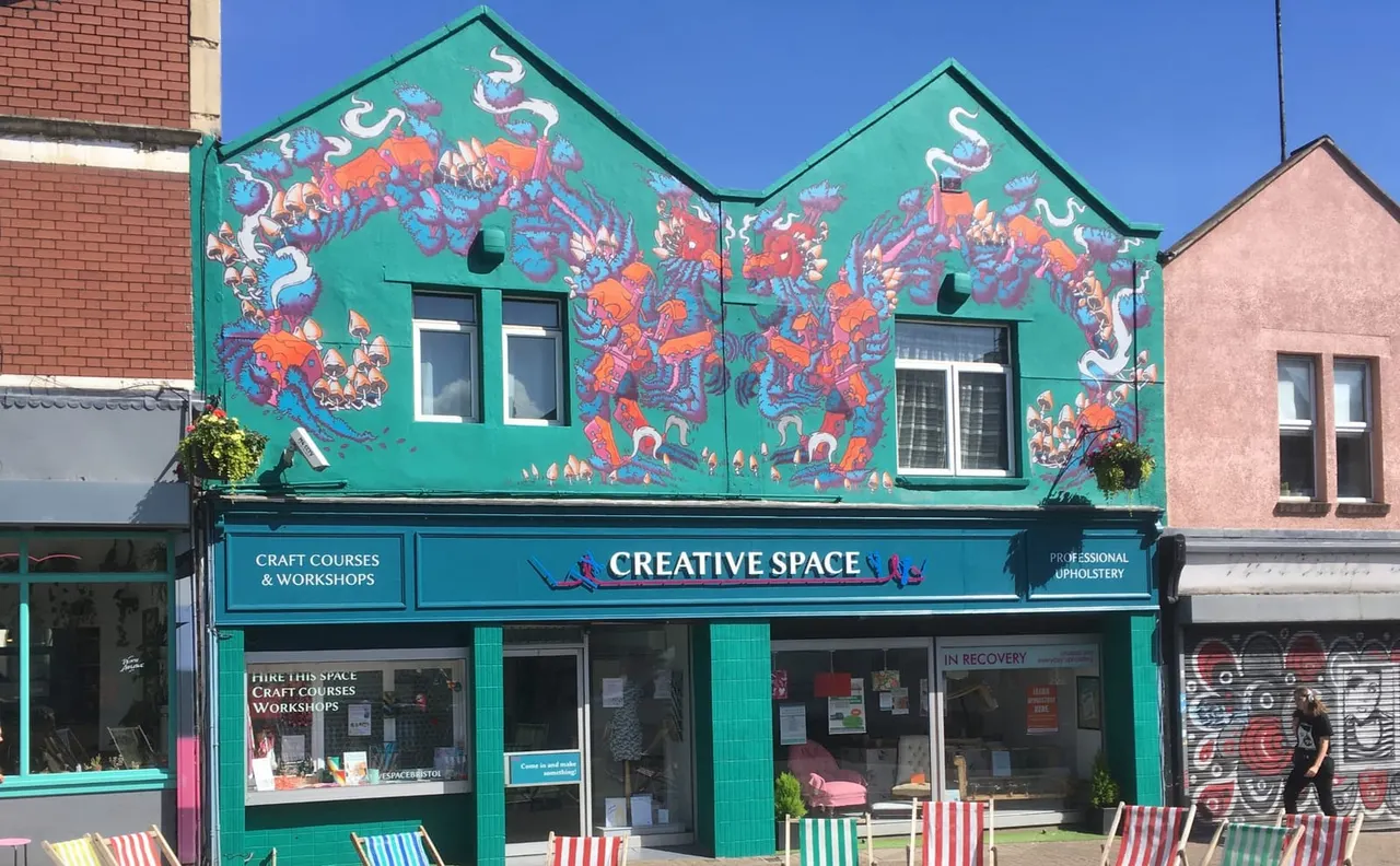 Turquoise storefront with colourful murals featuring dragons and mythical creatures, with deck chairs outside on a sunny day.