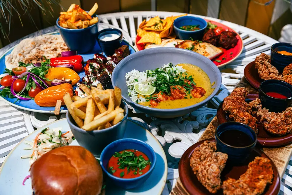 An overhead view of a table laden with colourful dishes including curry, fried chicken, chips, roasted vegetables, bread and dips.