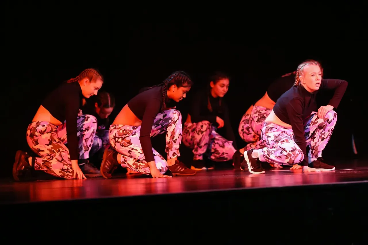 A group of young dancers performing a contemporary dance routine on stage under red stage lighting.