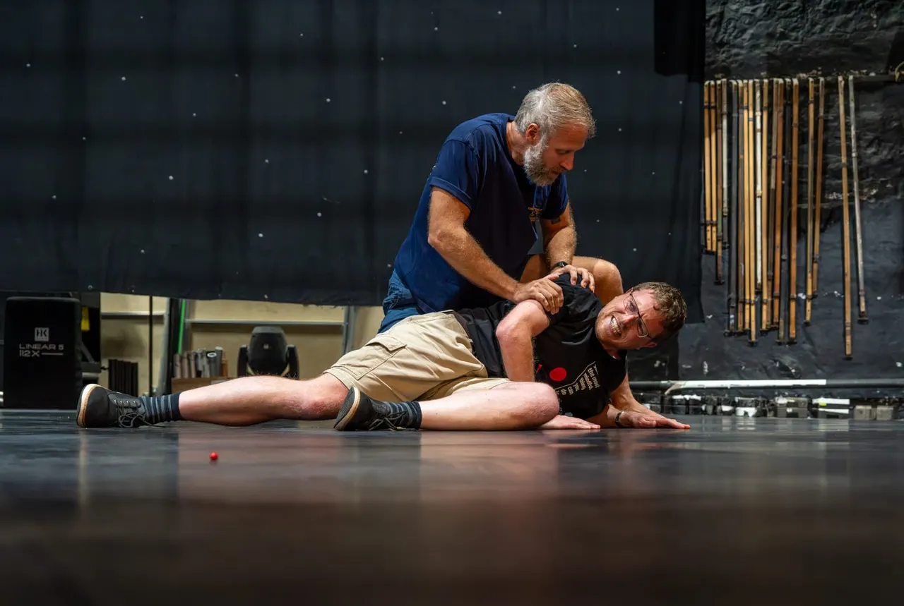 An instructor demonstrates a physical technique on a participant lying on a dark floor in a theatre space.