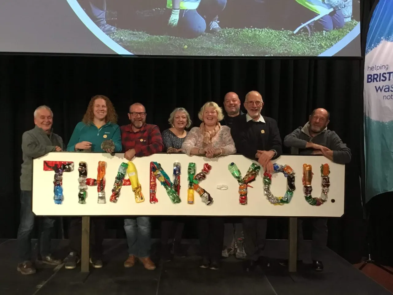 Eight people stand on stage holding a large sign spelling "THANK YOU" made from coloured recycled bottles.