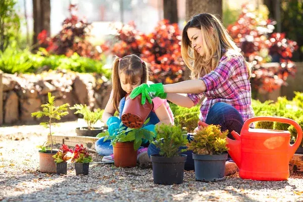 Woman and young girl planting flowers together in a sunny garden surrounded by potted plants and a red watering can.