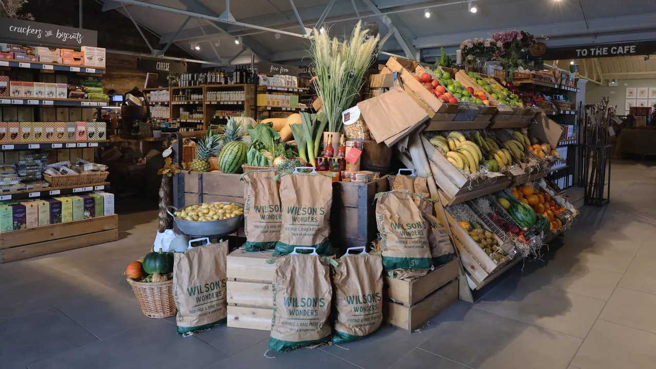 Farm shop interior with fresh produce display including fruit, vegetables, pineapples and bananas arranged in wooden crates and boxes.