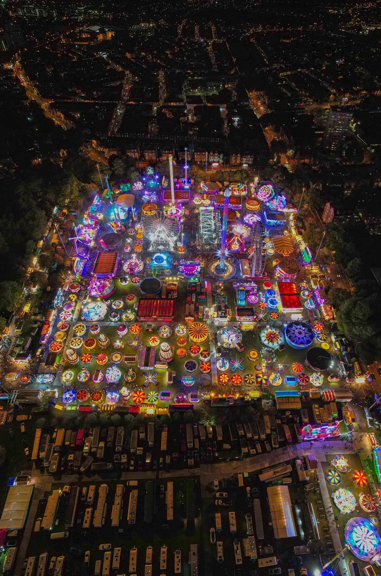 Aerial night view of a brightly lit fairground with colourful neon rides, attractions and a Ferris wheel surrounded by city streets.