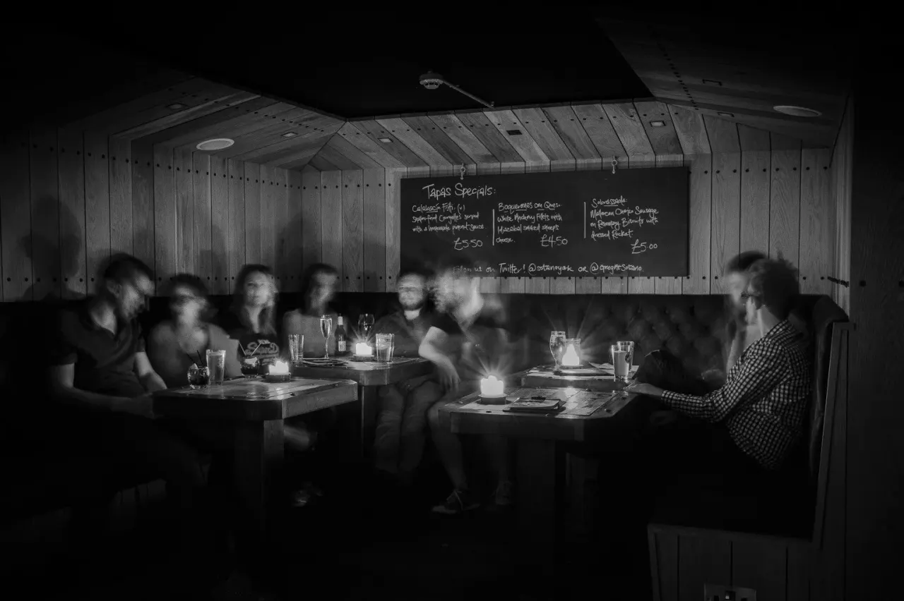 Group of diners seated at tables in a dimly lit wooden restaurant with a tapas menu board on the wall.