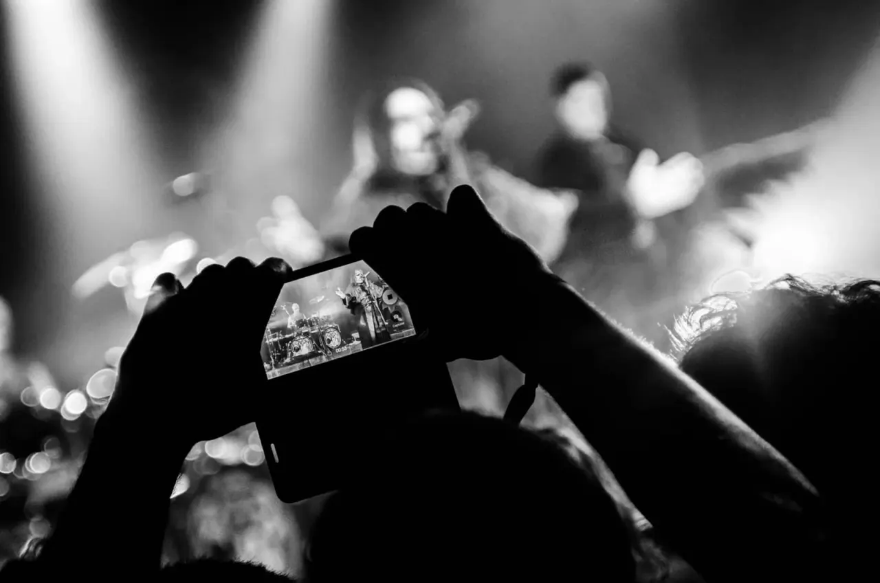 Person filming a live music performance on a smartphone, with performers and stage lights blurred in the background.
