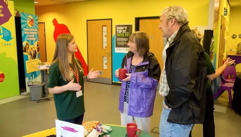 A staff member in a green shirt explains an exhibit to two visitors in a colourful indoor centre with educational displays on the walls.