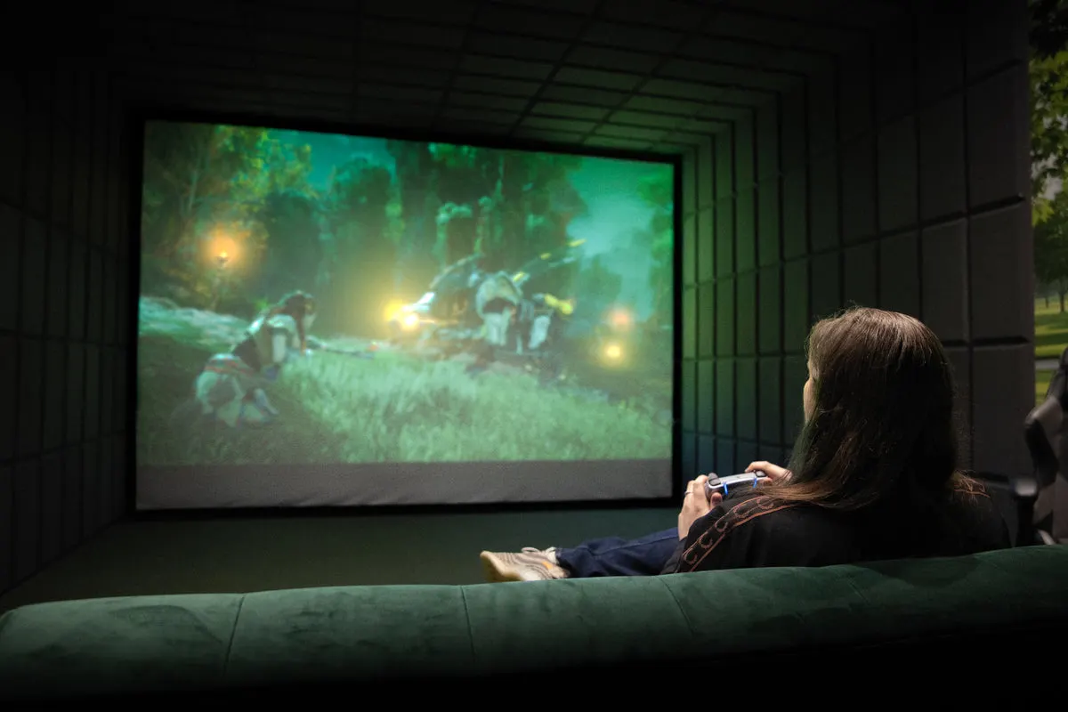 Person sitting on a green sofa watching a projected film scene with a game controller in hand.