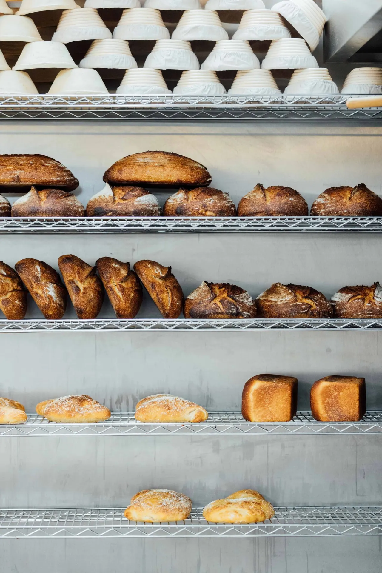 Shelves displaying fresh baked bread and rolls, including baguettes, round loaves, and square loaves, with white bowls stacked on top shelf.