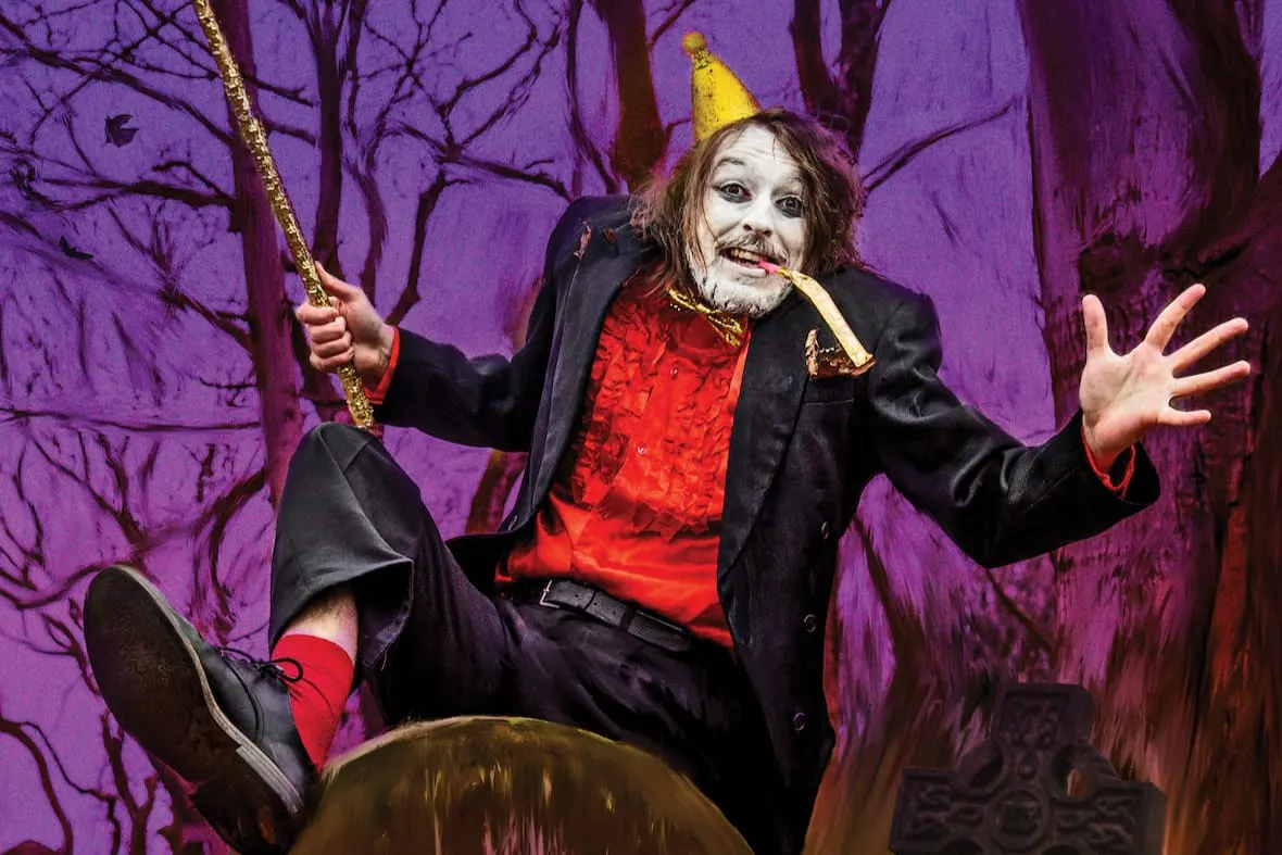 Performer in clown costume with white face paint, yellow hat and red outfit swinging on a rope against purple forest backdrop.