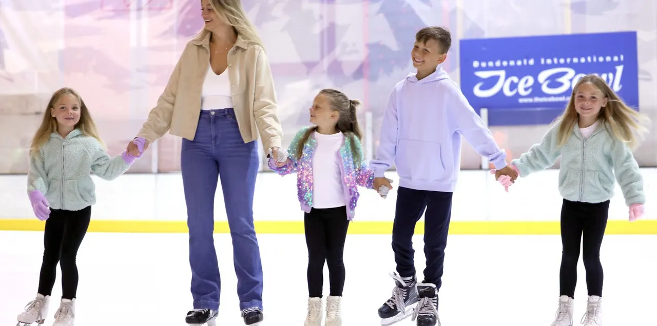 An adult and four children ice skating together on an indoor ice rink, holding hands and smiling.