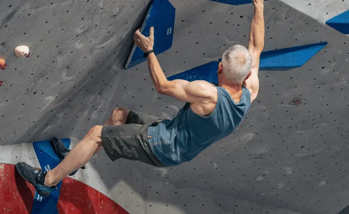 Man climbing an indoor rock wall with blue handholds and safety equipment.