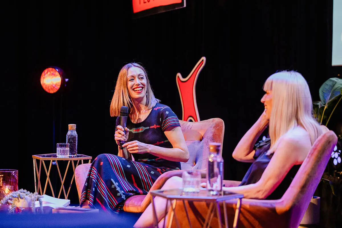 Two women on stage during a panel discussion or interview, with one holding a microphone, seated in front of neon signage and stage lighting.