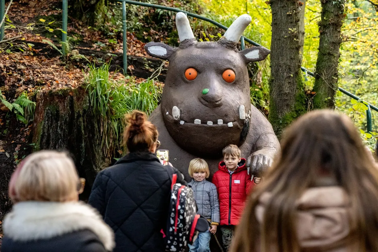 Visitors including young children posing for photos with a large cartoon monster sculpture in a woodland setting.
