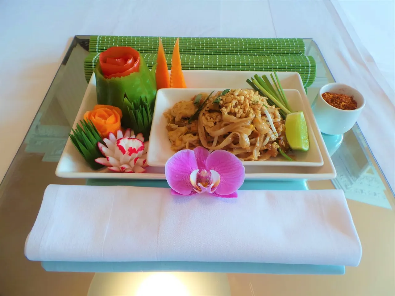 Pad Thai noodle dish served on white plate with decorative carved vegetables and fresh flowers, lime wedge and dipping sauce.