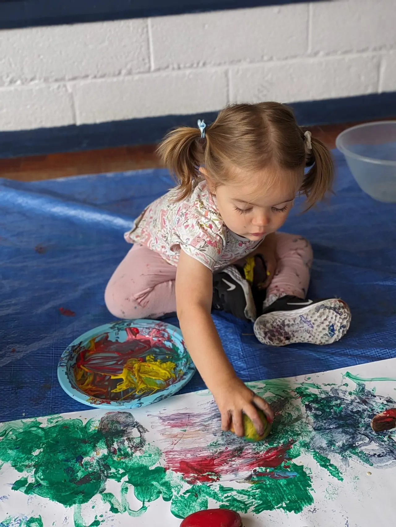 A young child sits on a blue mat, painting with colourful paints on paper whilst indoors.