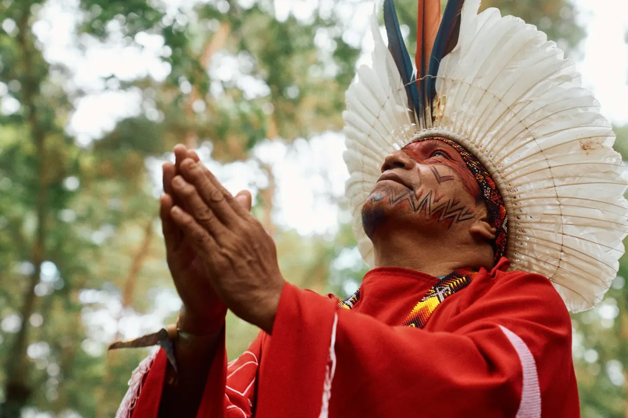Man wearing traditional indigenous headdress and red clothing with face paint, looking upward outdoors amongst trees.
