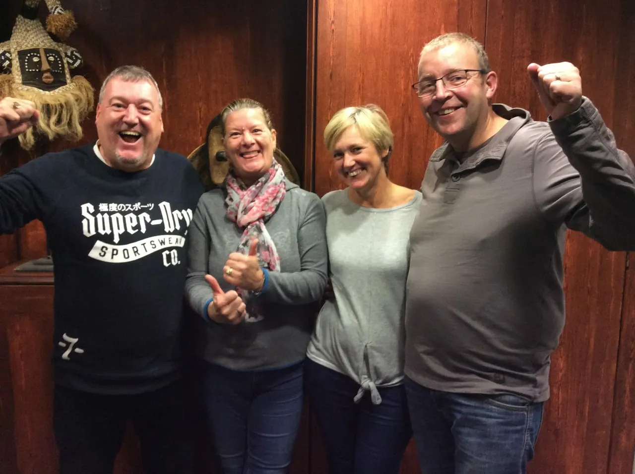 Four adults smiling and giving thumbs up inside a wood-panelled room with a decorated mask on the wall.