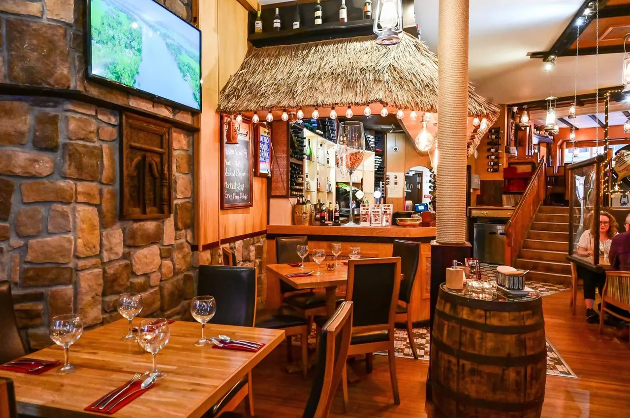 Interior of a tropical-themed restaurant with thatched roof bar, wooden tables set with glasses, and warm orange-toned walls.