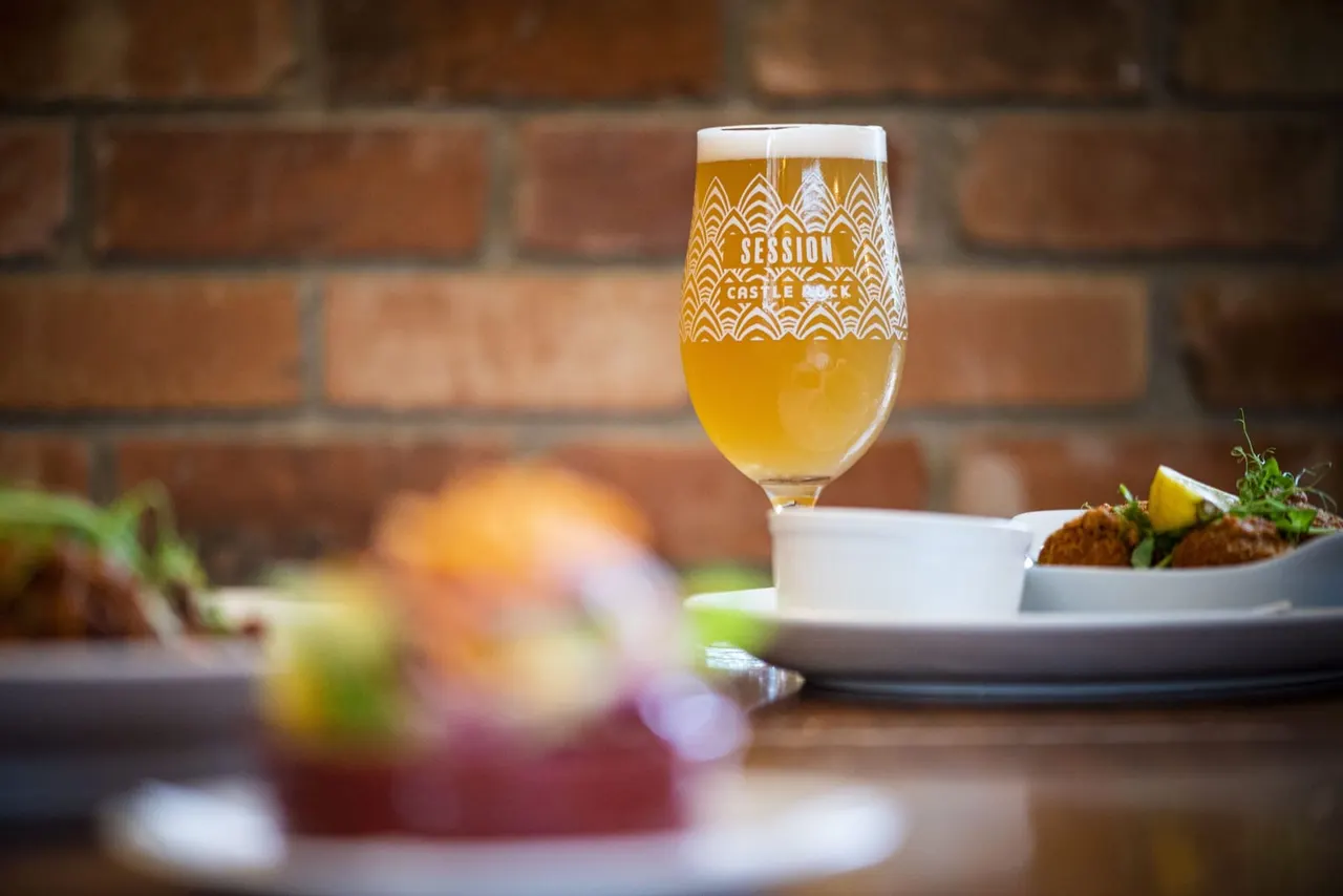 A glass of golden session ale sits on a table with food plates and a brick wall background.